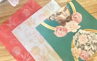 Three stacked notebooks with whimsical covers, featuring floral patterns and a classical portrait with a decorative halo on a wooden surface.
