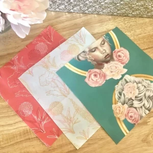 Three overlapping notebooks with artistic covers; one with red floral patterns, another with white botanical illustrations on pink, and one depicting a surreal portrait surrounded by flowers on green.