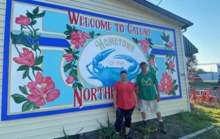 Two people standing proudly in front of a whimsical mural that reads "welcome to Callao, hometown of the northern neck," featuring vibrant flowers and a blue globe.