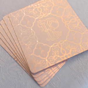 A stack of elegant copper-colored napkins with embossed floral patterns and a monogrammed initial "k" on a textured gray surface, evoking a whimsical aesthetic.