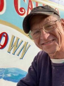 An elderly man with glasses and a cap smiles at the camera, standing in front of a whimsical mural with large painted letters and a blue wave design.