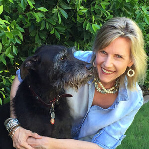 A woman with blonde hair smiling while embracing a large black dog in a Park West garden, surrounded by lush greenery. She wears large earrings and a pearl necklace.