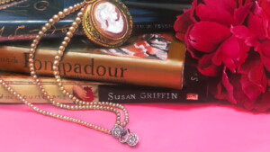A whimsical still life arrangement featuring a stack of books on Marie Antoinette and Madame de Pompadour, a pearl necklace with a cameo pendant, and a red rose, all resting on a