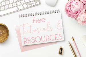 A desk with a notepad labeled "free tutorials & resources" in pink cursive, alongside a keyboard, a pink peony, pen, pencil, and whimsical decorative items in a clean,