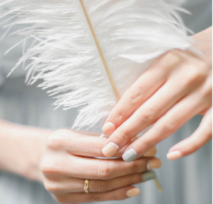 A close-up photo showcasing the delicate hands of a person holding a white feather, focusing on the whimsical engagement ring and beautifully manicured nails.