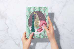A person's hands holding a Christmas card featuring a whimsical illustration of an animal dressed in a festive scarf, set against a colorful, surrealist background.