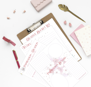 A neatly arranged desk with a clipboard holding a "creative bucket list" paper, surrounded by a pink notebook, gold pen, whimsical floral decorations, and office supplies on a white background.