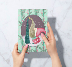 A person holds a holiday card featuring a surrealist illustration of a whimsical green Christmas tree with a gold star and pink decorations on a maroon backdrop, framed by a snowy scene and pine branches.