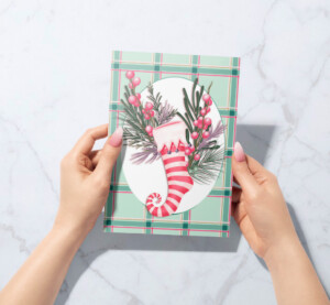 Hands holding a Christmas card featuring a festive plaid design with a surrealist painting of an elf’s leg and candy cane surrounded by pine branches and berries.