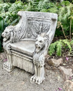 An ornate stone bench with lion sculptures on the arms, set against a background of lush ferns in Park West. The backrest features intricate carvings with a grapevine motif.