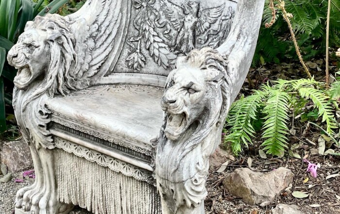 An ornate stone bench with lion sculptures on the arms, set against a background of lush ferns in Park West. The backrest features intricate carvings with a grapevine motif.