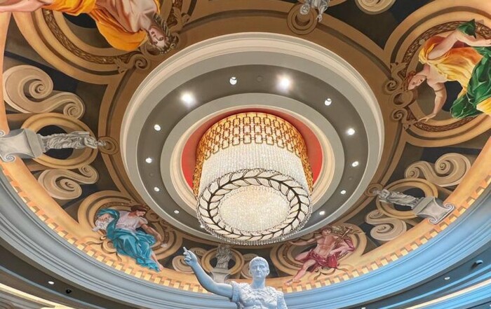 An ornate ceiling featuring swirling designs and classical figures in a rotunda, with a central glowing chandelier and a statue by Melanie Stimmell in the foreground.