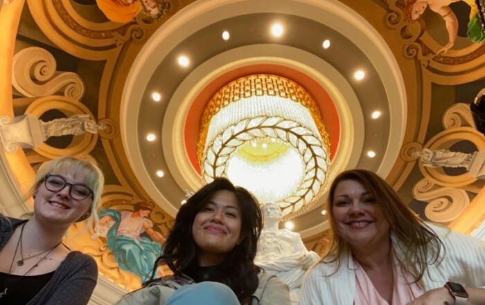 Three women smiling at the camera, sitting under Melanie Stimmell's ornate ceiling with a spiral design and a central chandelier, surrounded by mural artwork.