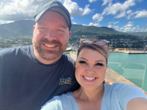 A smiling man and woman taking a selfie with a scenic view of a coastal town and hills in the background, under a clear blue sky at Park West.