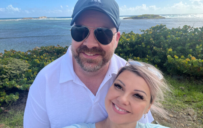 A smiling couple takes a surreal selfie with a scenic coastal background featuring lush greenery and a distant island under a bright blue sky. Both wear sunglasses; the man sports a cap.