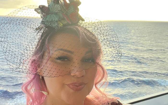 A woman with pink hair wearing a butterfly-adorned hat and a mesh veil smiles, posing on a ship deck at sunset over the ocean, reminiscent of a Melanie Stimmell painting.