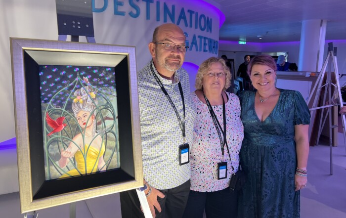 Three adults smiling next to a framed surrealist painting at an event with a "Park West" sign glowing in the background. Two women and one man are wearing event lanyards.
