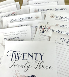 Printed motivational worksheets with inspirational phrases and headers, including "twenty twenty-three" in Melanie Stimmell's signature cursive style, spread out on a table.