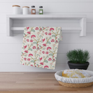 A floral-print kitchen towel hangs over a modern white shelf with decorative jars on top. Beside it, there's a whimsical potted plant and a wooden basket containing a loaf of bread, all