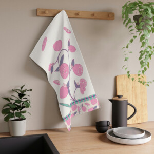 A kitchen towel with a whimsical pink and purple fruit design by Melanie Stimmell hangs on a wooden rack above a counter with a black coffee pot, cups, and plates, surrounded by potted