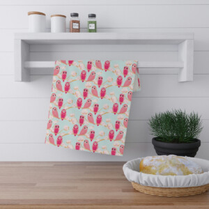 A kitchen towel with a pink and teal bird pattern hangs on a modern white shelf, accompanied by jars and a green plant, above a wooden countertop with a bread basket adorned with surrealism.