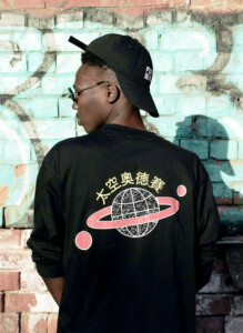 A young man wearing a black cap and a jacket with a whimsical design featuring Asian characters and a planet, stands in front of a graffiti-covered brick wall.