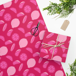 A whimsical Wrapping Paper Pink Lemons adorned with white citrus fruit slices decorates a tabletop, a matching gift box tied with a green string and a sprig of greenery beside it, accentuated by black.