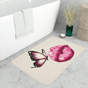 A bath mat featuring a whimsical pink heart and a pink and black butterfly design, placed on a bathroom floor next to a white bathtub and a wicker basket.