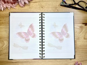 An open spiral notebook lies on a wooden table, displaying lined pages with soft pink butterfly illustrations. Pink flowers border the upper corners, a pair of eyeglasses sit on the top right corner, and a hand is partially visible in the bottom right corner.