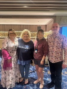 Four individuals are standing together indoors and smiling at the camera. The group consists of two women on the left, with one in a floral dress and the other in a Mickey Mouse shirt, and two people on the right, one in a maroon hoodie and the other in a floral button-up shirt.